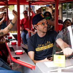 Driver steering a red pedal-powered party trolley through a downtown street while smiling passengers cheer and raise red cups