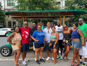 Ten friends in colorful summer outfits smiling and posing in front of a green-canopied open-air party trolley on a tree-lined city street.