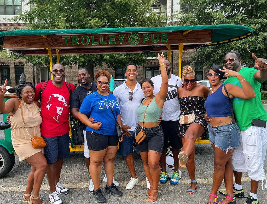 Cheerful group of ten adults posing on a green open-air party trolley during a sunny urban street tour, wearing colorful summer outfits and sandals, laughing and celebrating together.