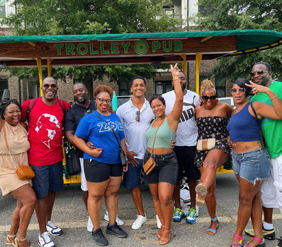 Cheerful group of ten adults posing on a green open-air party trolley during a sunny urban street tour, wearing colorful summer outfits and sandals, laughing and celebrating together.
