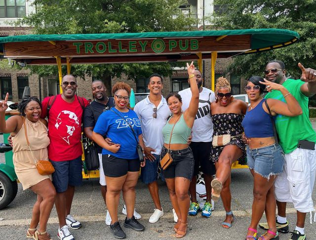 Cheerful group of ten adults posing on a green open-air party trolley during a sunny urban street tour, wearing colorful summer outfits and sandals, laughing and celebrating together.