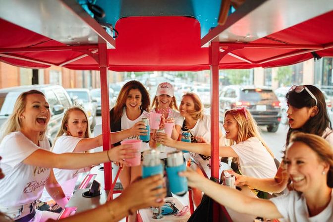 Group of women laughing and toasting drinks aboard a red pedal-powered party bike on a busy city street, lively urban celebration