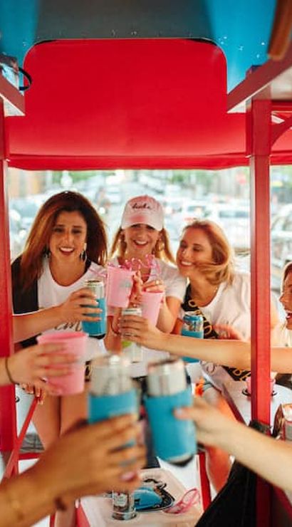 Group of women laughing and toasting drinks aboard a red pedal-powered party bike on a busy city street, lively urban celebration