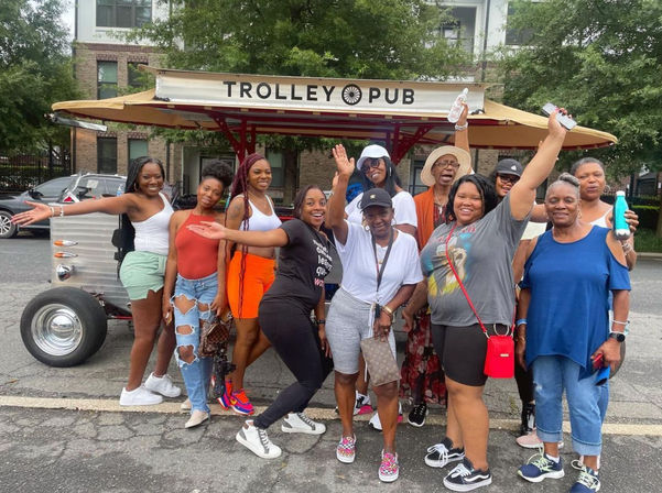 Group of smiling women posing and cheering in front of a parked open-air party trolley on a tree-lined city street, wearing casual summer outfits for a lively outing.