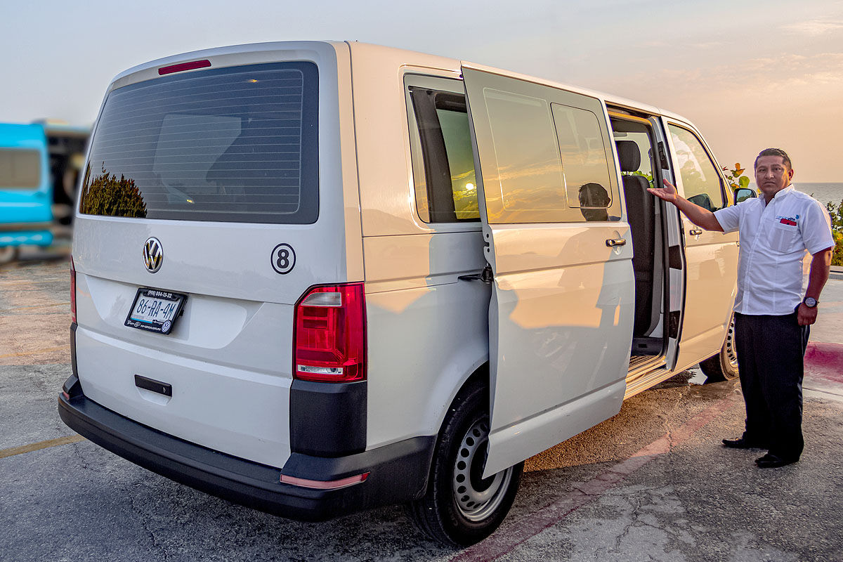 White Volkswagen passenger van with sliding side door open in a seaside parking lot at sunset, uniformed driver gesturing to invite passengers aboard.