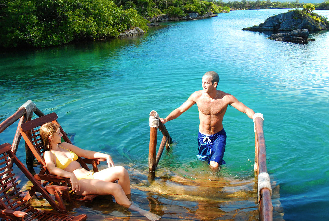 Sun-drenched tropical lagoon with clear turquoise water — a man in blue swim trunks climbs a wooden ladder while a woman in a yellow bikini relaxes on wooden lounge chairs near a mangrove-lined shore.
