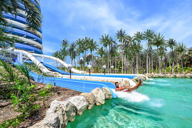 Man splashing into a turquoise lagoon from blue spiral water slides at a tropical resort water park with palm trees and a bright sunny sky