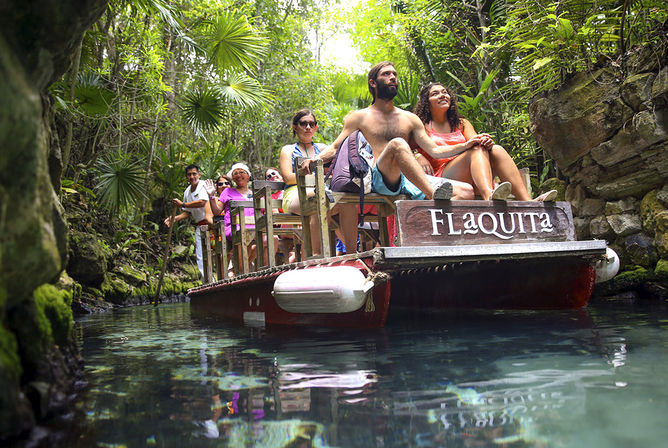 Group of people on a wooden raft-style boat enjoying a tropical cenote boat tour through a narrow jungle waterway with crystal-clear water, mossy rock walls and palms, guided by a pole.