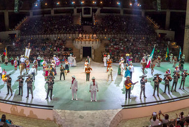 Vibrant Mexican folkloric show in a large amphitheater at night, mariachi band, dancers in colorful traditional costumes, flags, horses and a seated audience