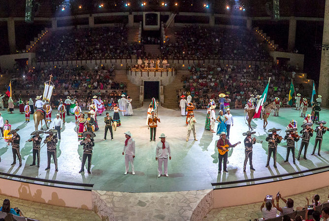 Vibrant Mexican folkloric show in a large amphitheater at night, mariachi band, dancers in colorful traditional costumes, flags, horses and a seated audience