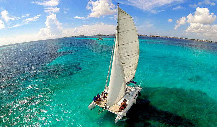 Aerial view of a white catamaran with billowing sail cruising over crystal-clear turquoise tropical water, passengers on deck, distant shoreline and puffy clouds