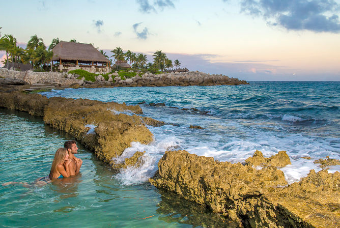 Romantic couple embracing in a turquoise tidal pool beside a rocky tropical shoreline with palm trees and a thatched-roof beach pavilion at sunset