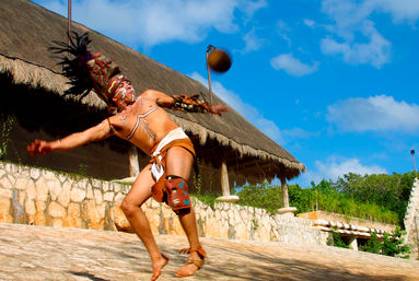 Costumed dancer wearing a feathered headdress and body paint tossing a ball on a sunlit stone plaza in front of a thatched-roof palapa under a bright blue sky.