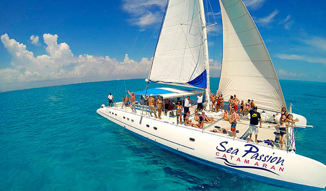 Sun-soaked group on a white sailing catamaran with full sails gliding over turquoise tropical waters under a bright blue sky