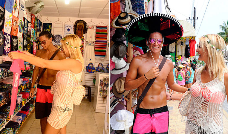 Two tourists shopping at a tropical beachside market: woman in a white crochet cover‑up browses colorful beachwear inside a souvenir shop while a shirtless man tries on a large sombrero and sunglasses by a stand of hats and t‑shirts.