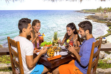Four friends enjoying colorful tropical cocktails and seafood at a wooden table under a thatched roof, seaside cliff with turquoise ocean and palm-fringed shoreline