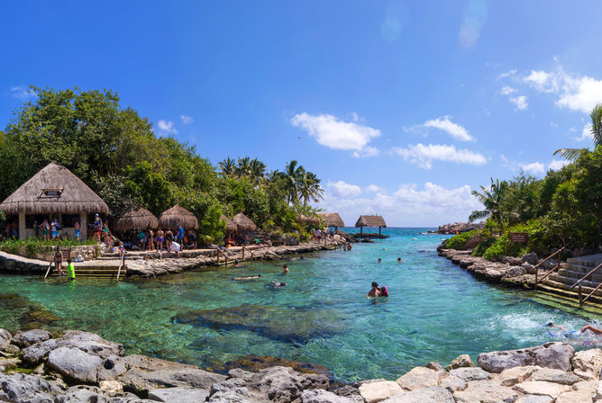 Sunny tropical lagoon with crystal-clear turquoise water, swimmers and snorkelers, thatched palapas and palm trees along a rocky shoreline.