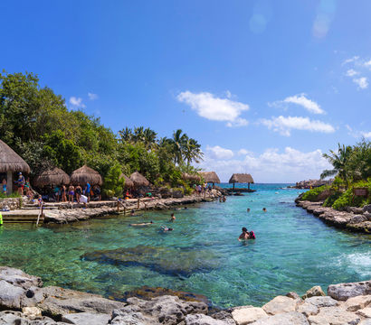 Sunny tropical lagoon with crystal-clear turquoise water, swimmers and snorkelers, thatched palapas and palm trees along a rocky shoreline.
