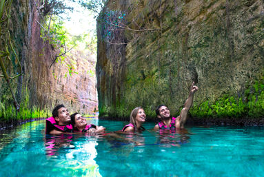 Four people in pink life jackets floating and smiling in clear turquoise water inside a narrow moss-covered limestone canyon, one pointing toward the sunlit rock walls and tree-lined opening — scenic canyon swim adventure.