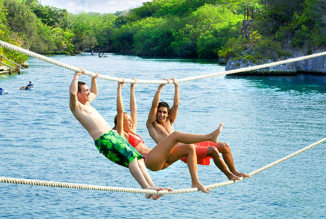 Three people in swimwear laughing as they hang from parallel ropes above a crystal-blue tropical lagoon with lush green shoreline