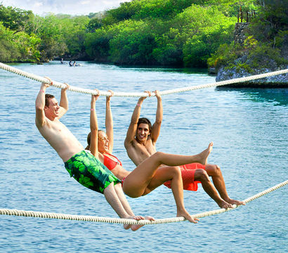 Three people in swimwear laughing as they hang from parallel ropes above a crystal-blue tropical lagoon with lush green shoreline