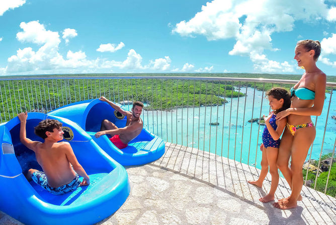 Smiling family at a tropical waterpark overlook — two riders in bright blue slide pods and a woman with a child by the railing, overlooking a turquoise lagoon, mangroves and a sunny sky.