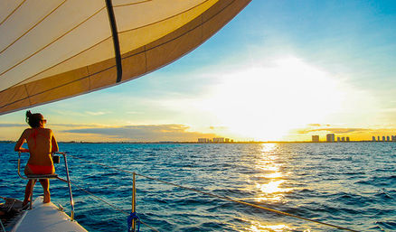 Person in a red swimsuit sitting on the bow of a sailboat at sunset, sailing toward a distant coastal skyline across shimmering blue ocean with a large sail overhead.