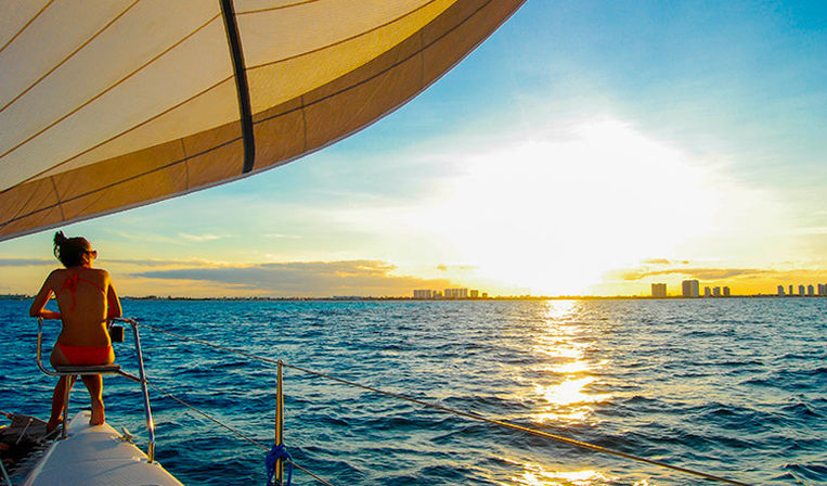 Person in a red swimsuit sitting on the bow of a sailboat at sunset, sailing toward a distant coastal skyline across shimmering blue ocean with a large sail overhead.