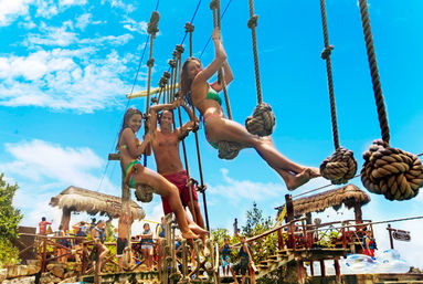 Three people in swimsuits swinging on a rope-obstacle course above a tropical waterpark with thatched huts, crowds below and a bright blue sky.