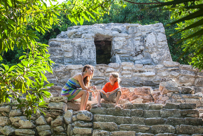 Woman and child chatting on ancient stone steps at a jungle ruin, framed by lush green foliage and sunlight
