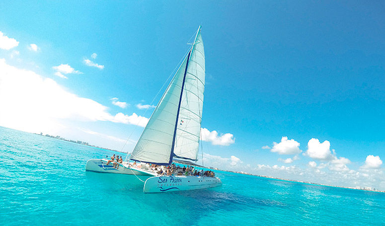 Catamaran with tall white sail cruising on turquoise tropical waters under a bright blue sky, passengers on deck and a distant shoreline on the horizon.
