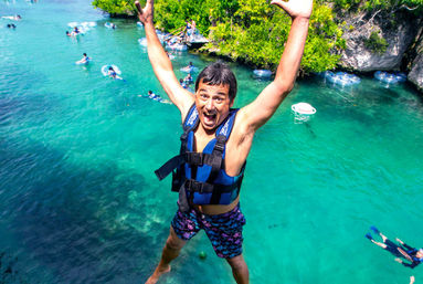Person in blue life jacket mid-air cliff jump over clear turquoise lagoon, with snorkelers, inner tubes, and green rocky shoreline — tropical adventure scene.