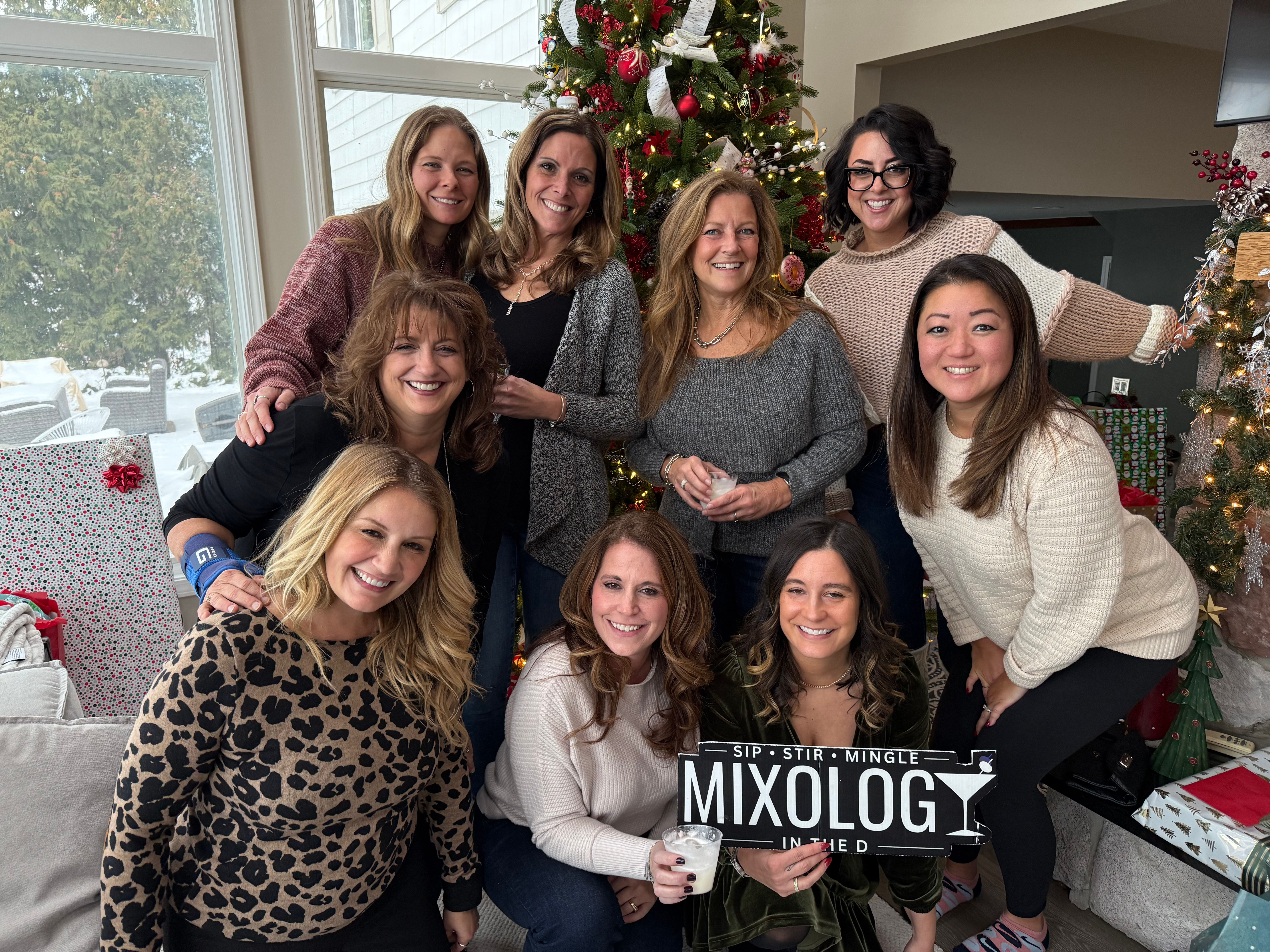 Group of smiling women at a cozy indoor holiday gathering by a decorated Christmas tree, holding a "Mixology" sign and drinks with a snowy yard visible through large windows.