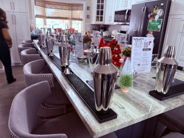 Modern kitchen island turned cocktail bar with marble countertop, stainless steel cocktail shakers on rubber mats, gray nailhead barstools and small holiday decorations