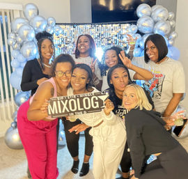 Eight women posing at a home mixology party, holding a 'MIXOLOGY' sign and drinks in front of a silver balloon arch and sequin backdrop.