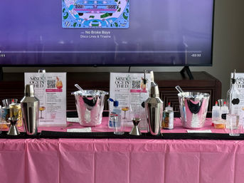 Home mixology station on a pink tablecloth with metal cocktail shakers, jiggers, ice buckets, squeeze bottles and mixers arranged on a bar mat in front of a TV showing colorful album art — DIY cocktail party setup.