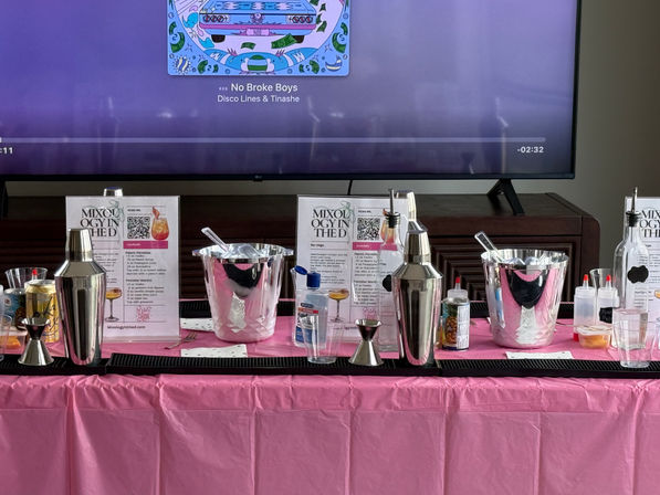 Home mixology station on a pink tablecloth with metal cocktail shakers, jiggers, ice buckets, squeeze bottles and mixers arranged on a bar mat in front of a TV showing colorful album art — DIY cocktail party setup.