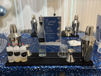 Indoor cocktail station at a party: stainless-steel shakers and jiggers, squeeze bottles of mixers, and a clear liquor bottle on a black bar mat over a blue confetti tablecloth with a cocktail menu and balloons in the background.