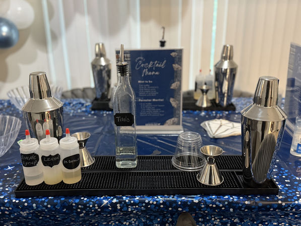 Indoor cocktail station at a party: stainless-steel shakers and jiggers, squeeze bottles of mixers, and a clear liquor bottle on a black bar mat over a blue confetti tablecloth with a cocktail menu and balloons in the background.