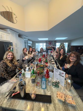 Smiling group of women raising cocktails around a marble kitchen island at a festive holiday cocktail party, with bottles, shakers, garnishes and mini Christmas trees on the counter and a decorated living room with a Christmas tree in the background.