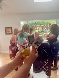 At-home bachelorette party — hands hold a 'Bachelorette Bash' glass and two instant photos while guests chat by a window with rose-gold 'BRIDE' balloons and a green backyard visible.