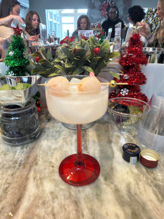 Festive lychee cocktail in a red-stemmed coupe on a marble countertop, garnished with two lychees on a skewer, surrounded by mini glittery Christmas trees and holiday bar decor with people blurred in the background.