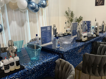 Indoor mixology station for a cocktail class with blue sequin tablecloths, cocktail shakers, syrup squeeze bottles, clear ice bowls, instruction cards, hand sanitizer and a white-and-blue balloon garland.