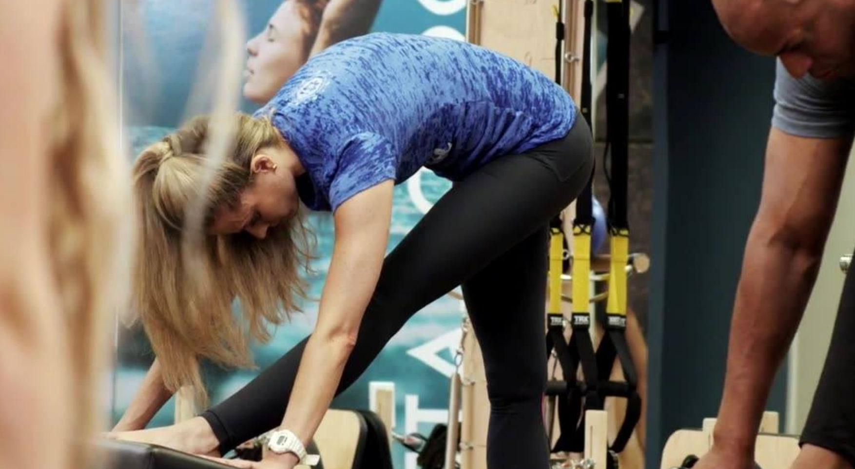 Woman in a blue shirt bending forward and stretching on a Pilates reformer in an indoor fitness studio with TRX straps and a trainer nearby.