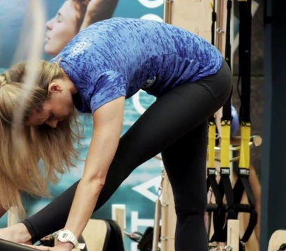 Woman in a blue shirt bending forward and stretching on a Pilates reformer in an indoor fitness studio with TRX straps and a trainer nearby.
