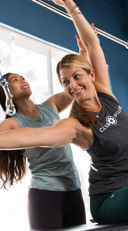 Two women in a bright Pilates studio practicing on a Cadillac reformer; an instructor assists a client with a seated side stretch using metal bars, springs, straps and sheepskin loops.