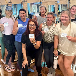 Smiling group of women in an indoor Pilates/fitness studio holding name cards after class, wearing workout clothes in front of reformer equipment and mirrors.