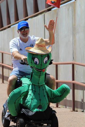 Smiling man in a blue cap and sunglasses rides a motorized scooter adorned with a large green cactus costume wearing a straw sombrero, waving on a sunlit outdoor path.
