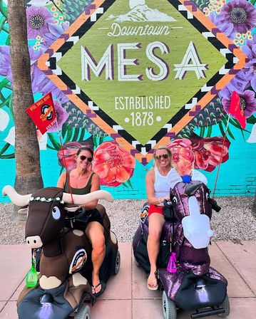 Two smiling women on novelty motorized bull scooters pose in front of a vibrant floral mural reading “Downtown MESA Established 1878” in Mesa, Arizona.