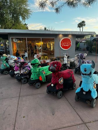 Colorful mobility scooters dressed as stuffed animals and monsters lined up outside an ice cream shop storefront at dusk with palm trees overhead